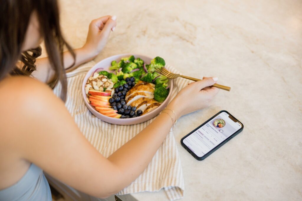 woman sitting in front of a plate of chicken, veggies, and fruits with a phone off to the side showing the cysterhood app recipe page