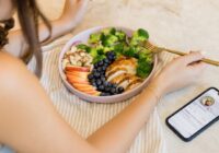 woman sitting in front of a plate of chicken, veggies, and fruits with a phone off to the side showing the cysterhood app recipe page